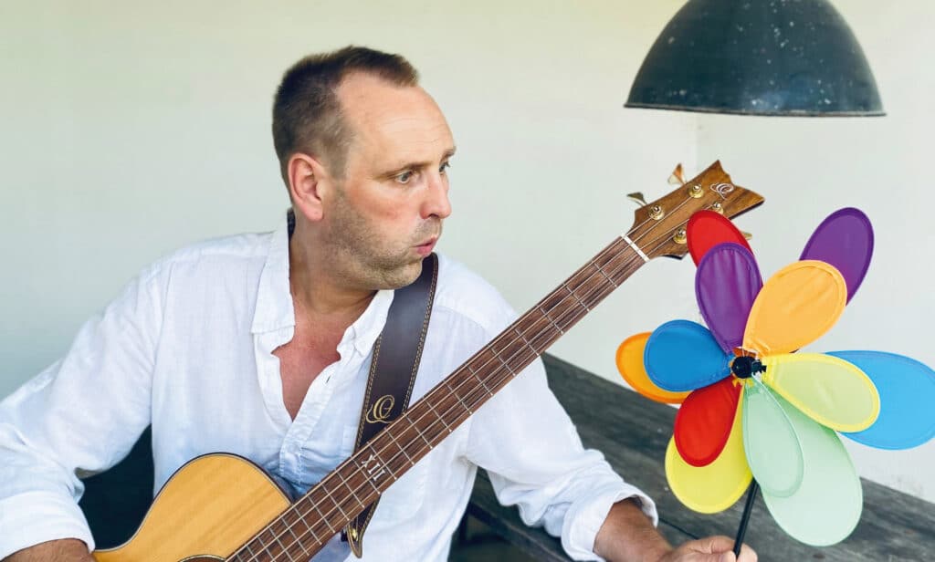 Musiker Tom Brückner hält eine Gitarre und eine bunte Blumenfigur vor einer hellen Innenraumwand.