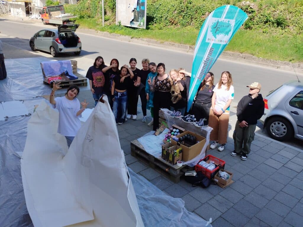 Gruppe junger Menschen an einem Straßenstand mit Bastelmaterialien, großem Papierboot und türkisfarbenem Banner; im Hintergrund eine Straße und begrünte Böschung