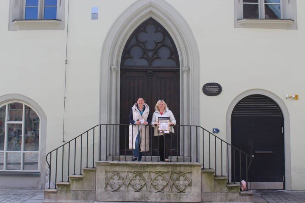 Märchenstunde „Der kleine Topf“ in Deggendorf Zwei Frauen stehen mit einem gerahmten Plakat vor einem spitzbogigen Eingang eines historischen Gebäudes in Deggendorf.