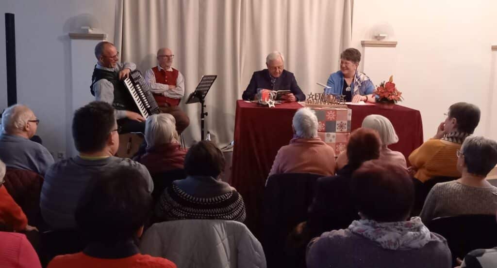 Publikum verfolgt eine Adventsveranstaltung mit Akkordeonmusik und einer Gesprächsrunde an einem dekorierten Tisch.