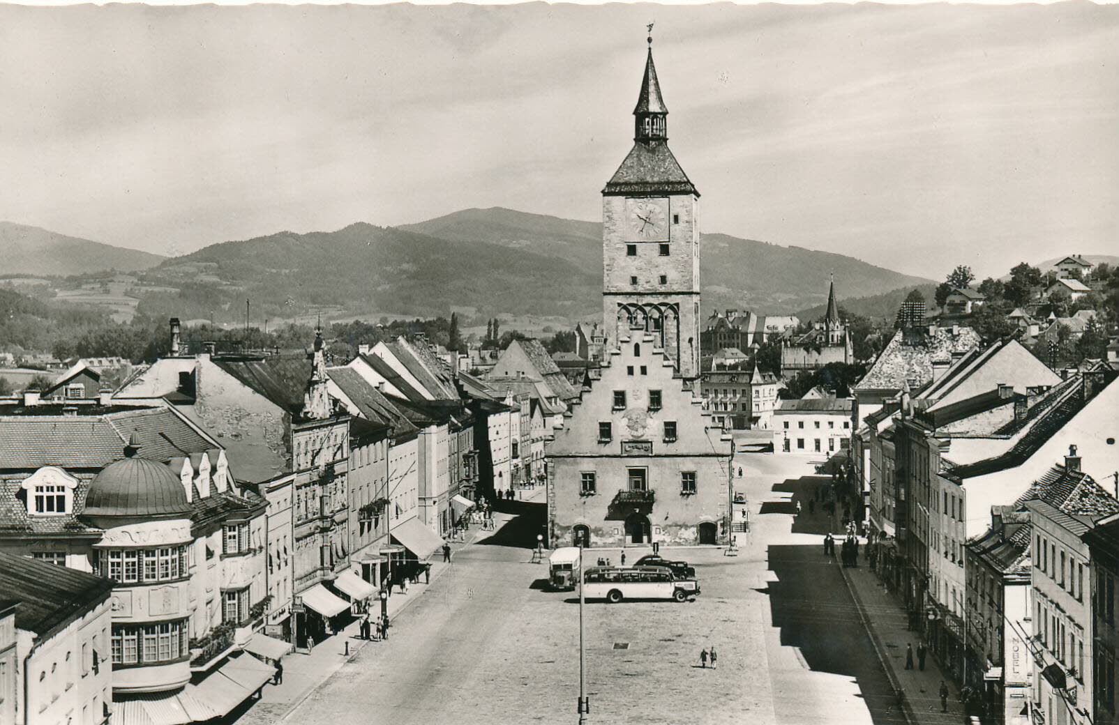 Schwarzweißaufnahme einer breiten Straße mit historischen Häusern und einem markanten Turm im Zentrum; im Hintergrund liegen bewaldete Hügel.