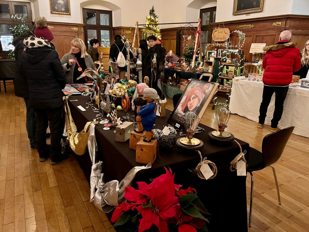 Besucherinnen und Besucher stöbern an Ständen mit handgefertigtem Schmuck, Dekorationen und Geschenkartikeln in einem weihnachtlich geschmückten Saal.
