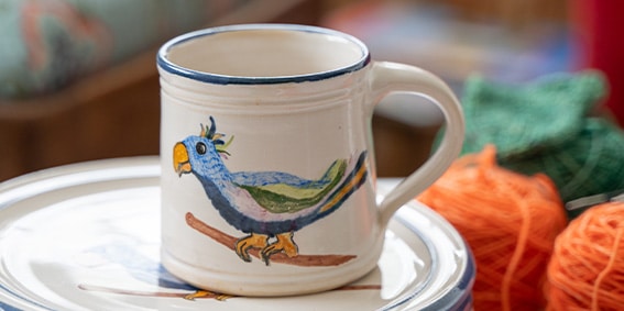 Strickcafé: Tasse mit handgemaltem Vogelmotiv Weiße Tasse mit blauem Rand und handgemaltem Vogelmotiv auf einem Teller; daneben liegen orangefarbenes und grünes Garn.