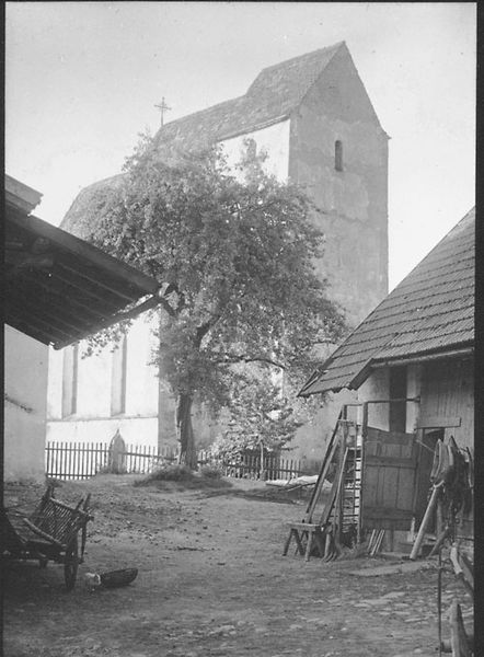 Schwarzweißaufnahme einer historischen Dorfstraße mit der Pfarrkirche von Schaching im Hintergrund, umgeben von Bäumen und landwirtschaftlichen Gebäuden.