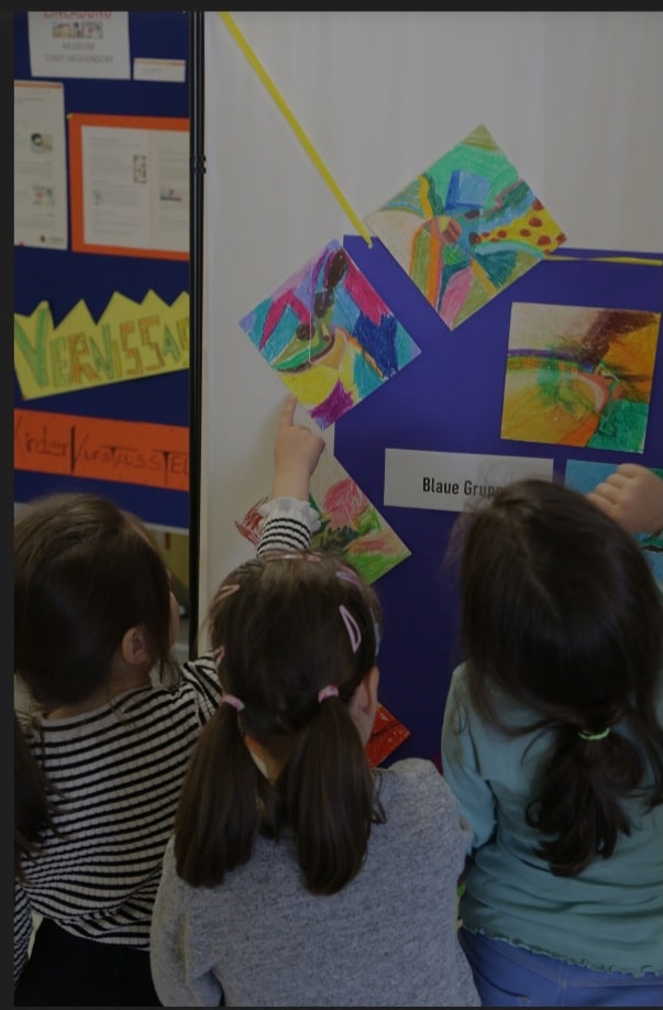 Kinder betrachten und gestalten farbenfrohe Kunstwerke an einer Ausstellungswand.