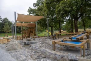 Wasserspielplatz mit Holzspielgeräten, Wasserrinnen, Sandflächen und Sonnensegeln in einer Parkanlage