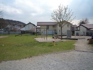 Grünfläche mit kleinem Spielplatz, jungen Bäumen und Wohnhäusern vor bewaldeten Hügeln