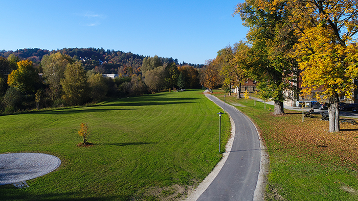 Asphaltierter Gehweg führt durch eine weitläufige, herbstlich gefärbte Parklandschaft mit Rasenflächen und Bäumen.