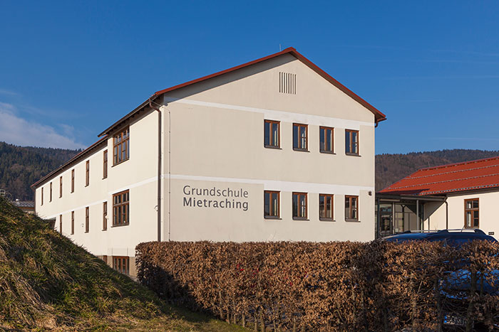 Gebäude der Grundschule Mietraching mit beschrifteter Fassade, Hecke und umliegender bewaldeter Hügellandschaft bei klarem Himmel