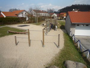 Gepflasterter und geschotterter Außenbereich mit niedrigen Holzpfosten, Sitzgelegenheiten, einem Kletterelement und Wohnhäusern im Hintergrund.