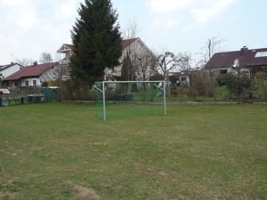 Grasfläche mit kleinem Fußballtor auf einem Bolzplatz in Rettenbach; dahinter stehen Wohnhäuser und Bäume.