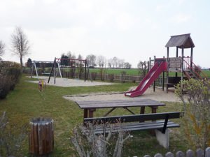 Ländlicher Spielplatz mit Rutsche, Schaukeln, Kletterturm und Sitzbank auf einer Grünfläche