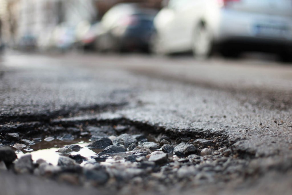 Nahaufnahme eines Schlaglochs mit losem Asphalt und einer Pfütze auf einer Straße; unscharfe Fahrzeuge stehen im Hintergrund.