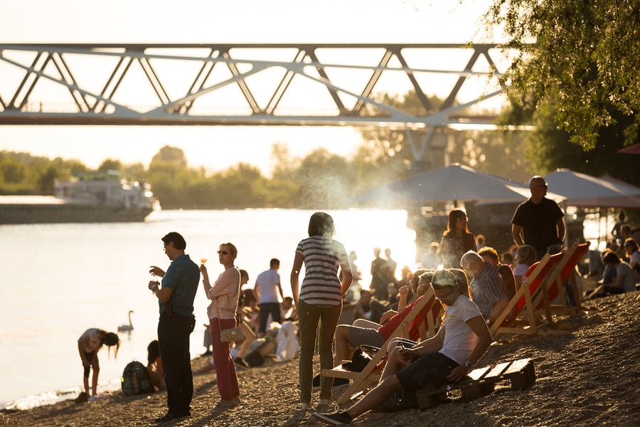 Sommerabend am Rhein in Dormagen Menschen sitzen und stehen an einem belebten Rheinufer im Abendlicht; im Hintergrund sind eine Brücke und das gegenüberliegende Ufer zu sehen.