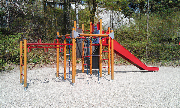 Leerer Spielplatz mit bunter Kletteranlage, Hängebrücke und roter Rutsche auf hellem Kies zwischen Bäumen