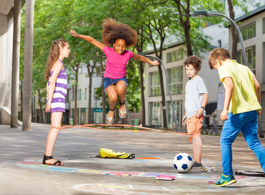 Vier Kinder spielen auf einem begrünten Platz mit einem Springseil, Straßenkreide und einem Fußball.