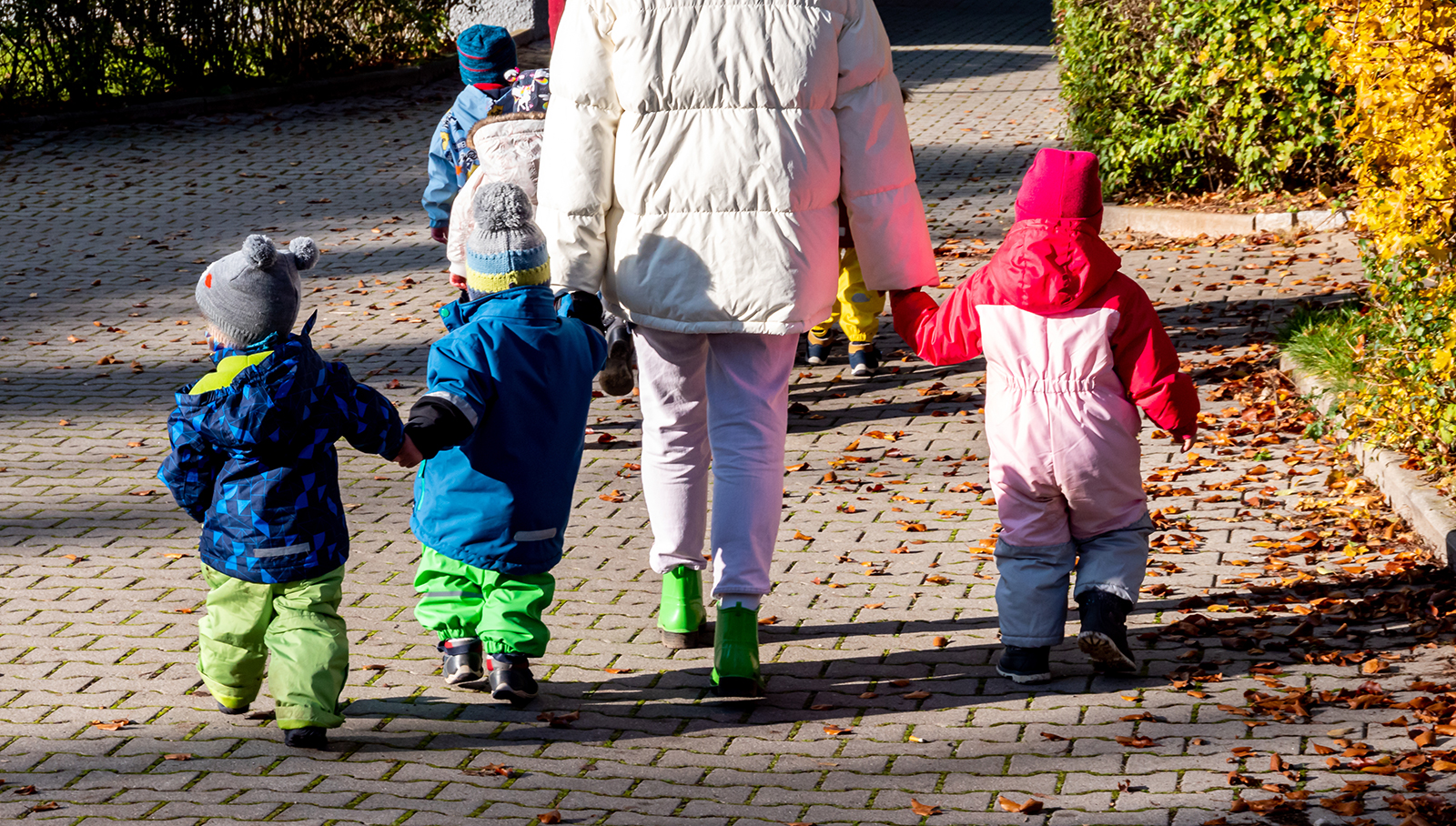 Eine erwachsene Person geht mit vier Kindern in bunten Winterkleidern über einen gepflasterten Weg.