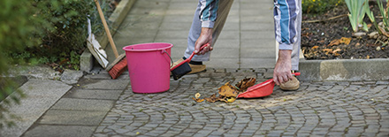 Person kehrt mit einem Besen Laub von einem gepflasterten Gehweg in eine rote Kehrschaufel.