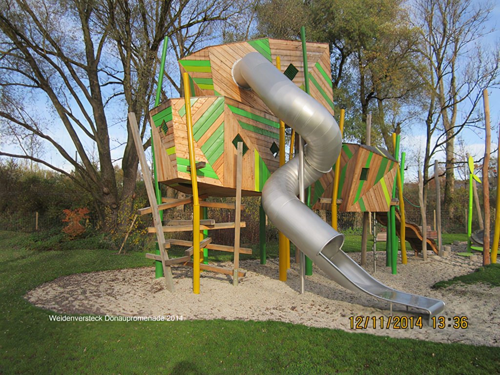 Kreativer Holzspielplatz mit großer silberner Röhrenrutsche, Kletterelementen und farbigen Holzwänden im Grünen
