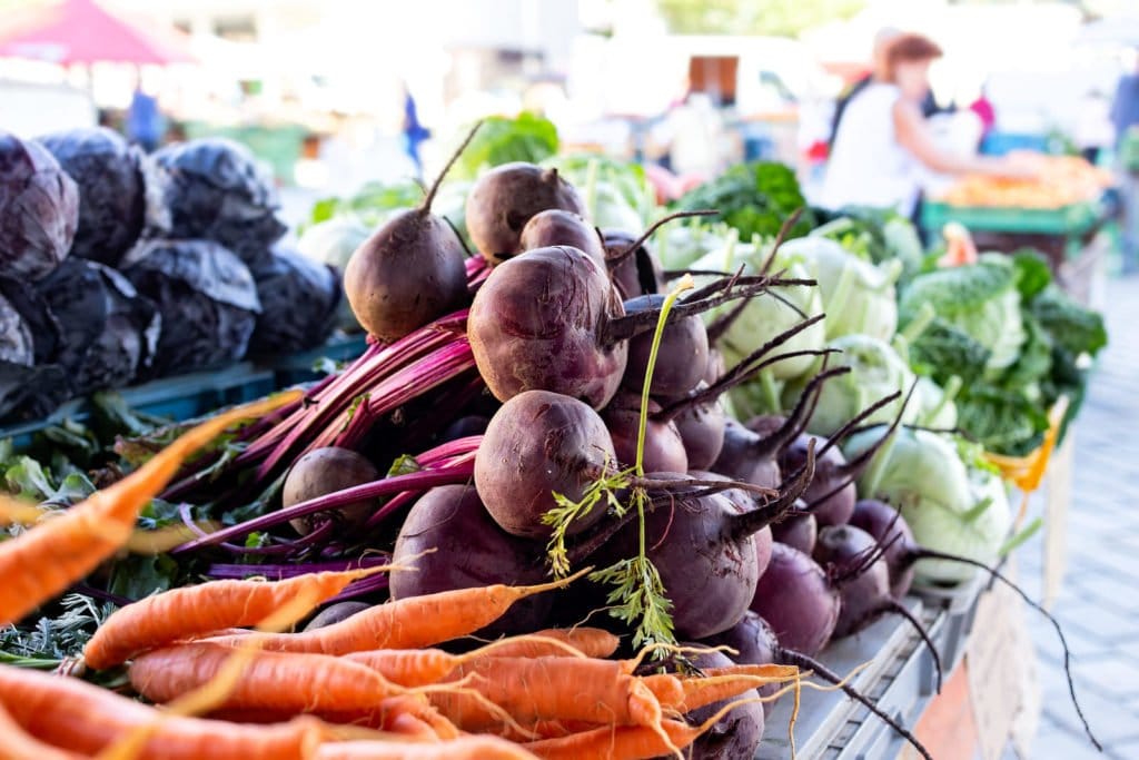 Bund Rote Bete und Möhren liegen auf einem Marktstand; dahinter ist weiteres frisches Gemüse zu sehen.