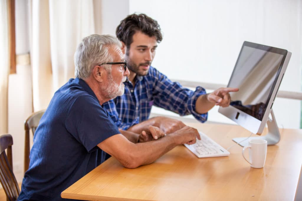 Beratung zwischen den Generationen am Computer Älterer Mann und jüngerer Mann betrachten gemeinsam einen Computerbildschirm am Tisch.