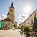 Stadtansicht mit Kirche am Marktplatz Historische Kirche mit Uhrturm an einem sonnigen Marktplatz; rechts stehen bepflanzte Außentische vor Häusern.