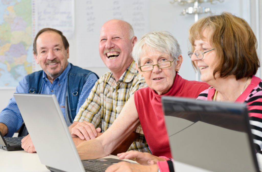 Vier ältere Erwachsene sitzen gemeinsam an Laptops und lächeln in einem hellen Raum.