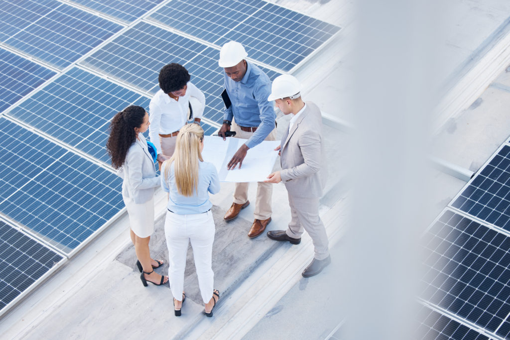 Fünf Personen besprechen Pläne auf einem Dach zwischen großflächigen Solarmodulen.