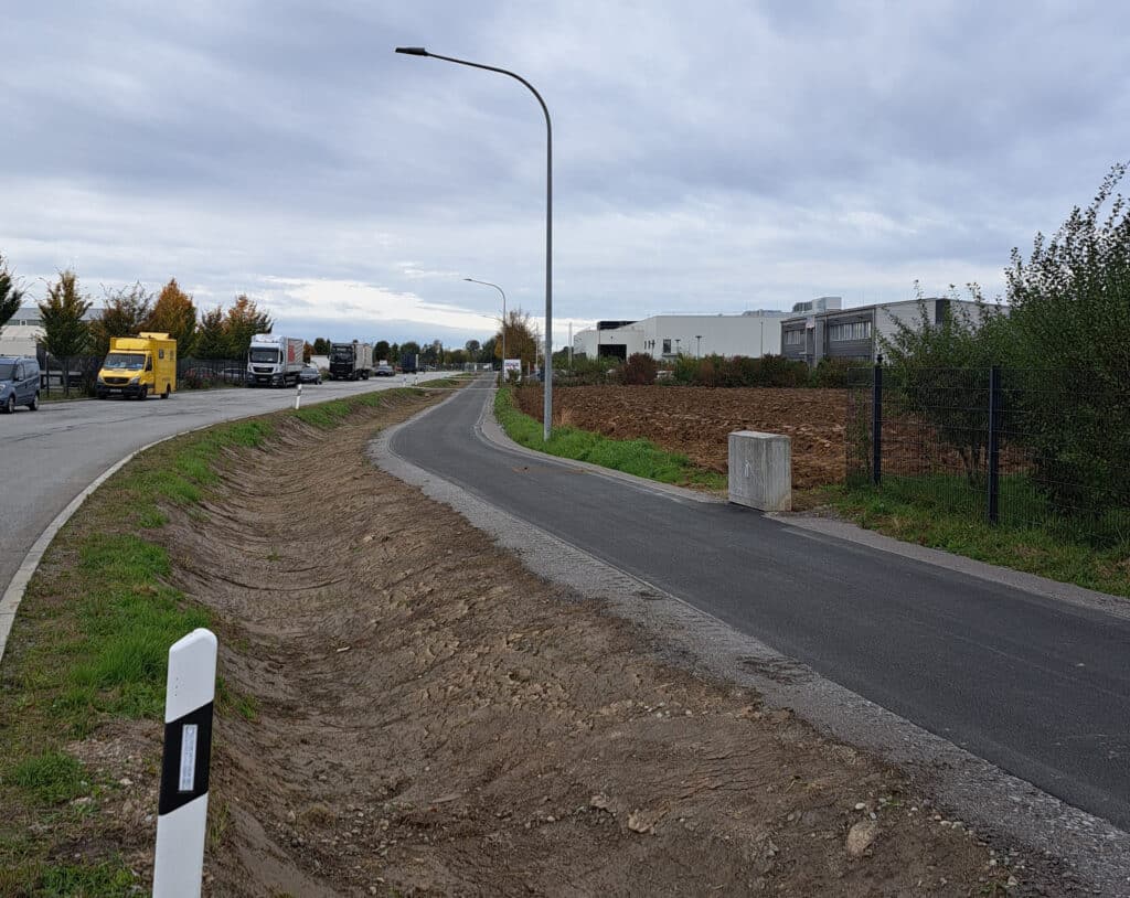 Geh- und Radweg an einer Straße Neu angelegter Geh- und Radweg neben einer Straße mit Straßenlaterne, Leitpfosten und Gewerbegebäuden unter bewölktem Himmel.