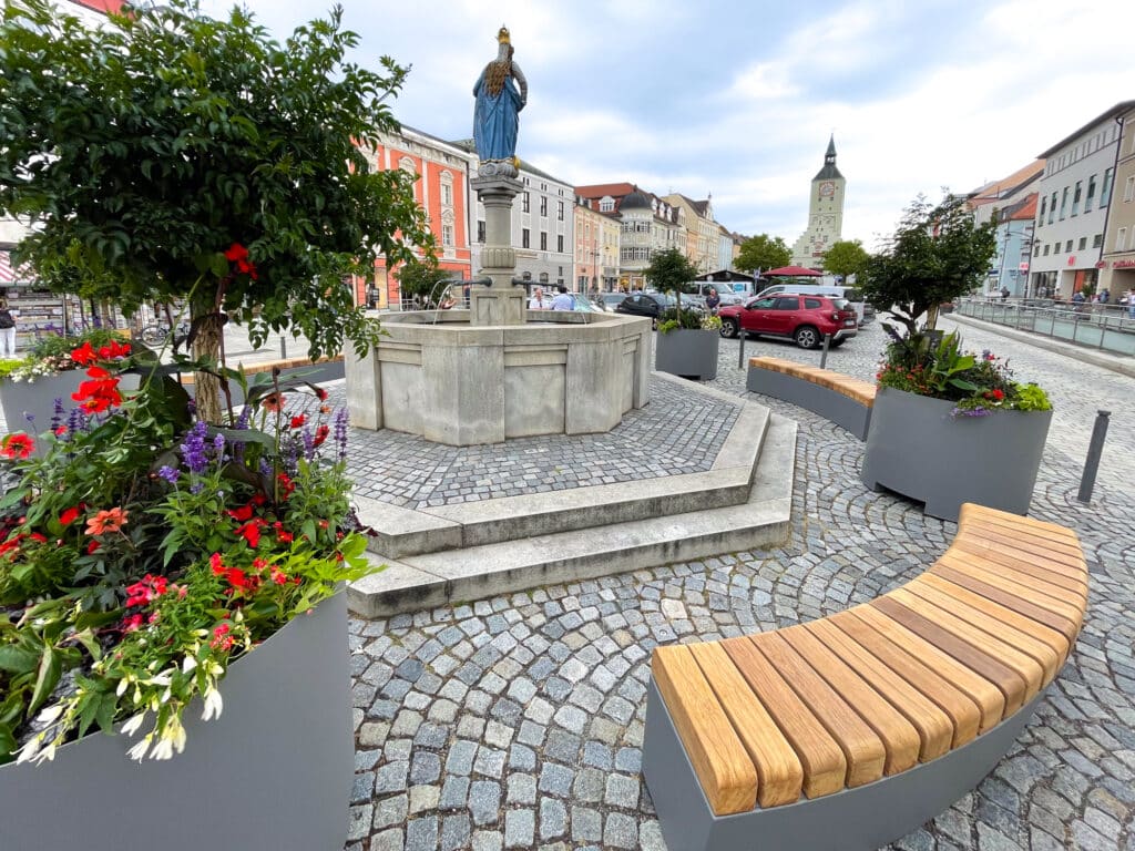 Geschwungene Holzbank und bepflanzte Pflanzkübel am Luitpoldplatz vor einer Marienstatue und dem Stadtturm