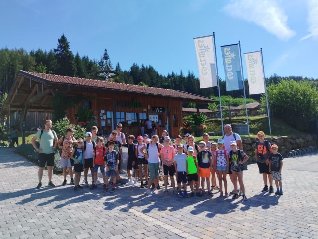 Gruppe aus Kindern, Jugendlichen und Betreuungspersonen steht bei sonnigem Wetter vor einer bewaldeten Ausflugsgaststätte mit Fahnen und Holzhütte.