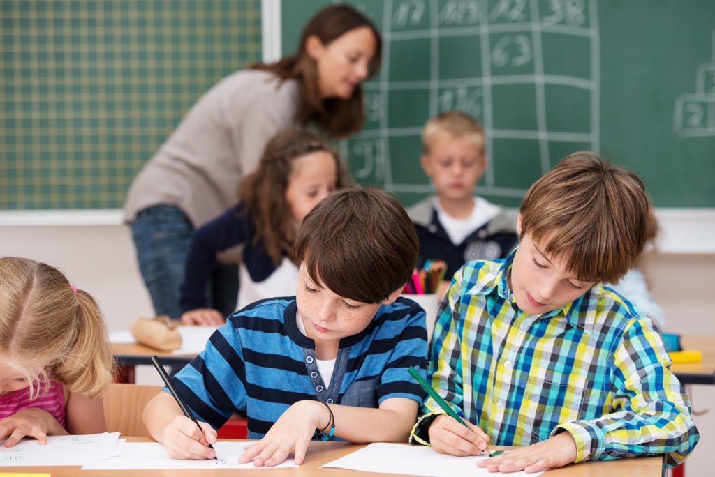 Mehrere Kinder malen und schreiben an einem Tisch, während eine Betreuerin und weitere Kinder im Hintergrund vor einer Tafel stehen.