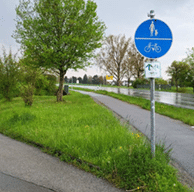 Blaues Verkehrszeichen für einen gemeinsamen Geh- und Radweg neben einer Straße mit Bäumen und Wiesen