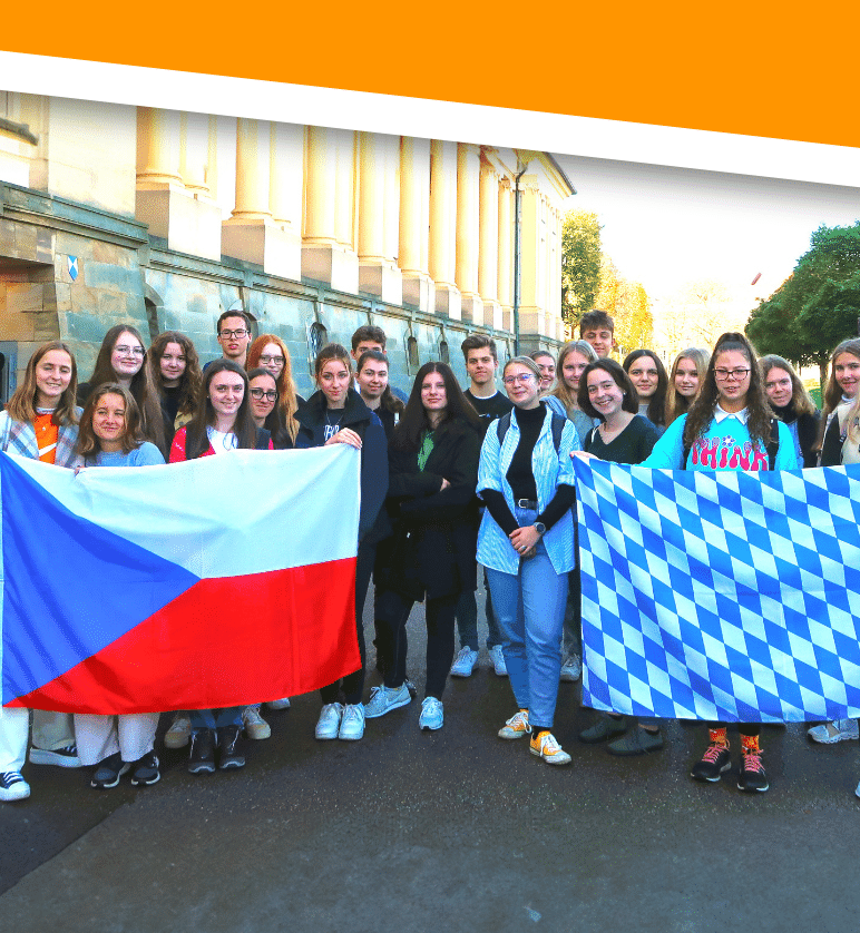 Gastfamilien gesucht: Jugendliche mit tschechischer und bayerischer Flagge Gruppe Jugendlicher hält vor einem historischen Gebäude eine tschechische Flagge und eine blau-weiß karierte bayerische Flagge.