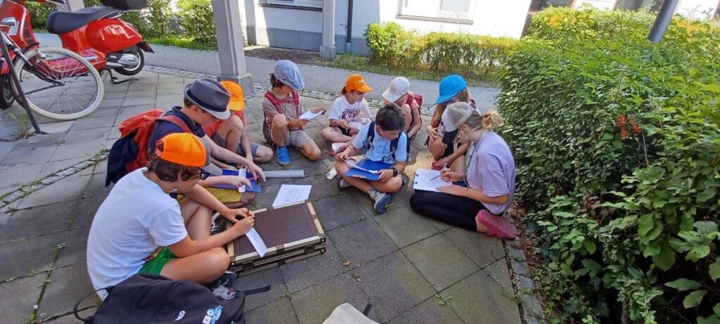 Eine Gruppe von Kindern und Jugendlichen sitzt mit Schreibunterlagen und Bastelmaterial auf einem gepflasterten Platz neben Fahrrädern und einem Motorroller.