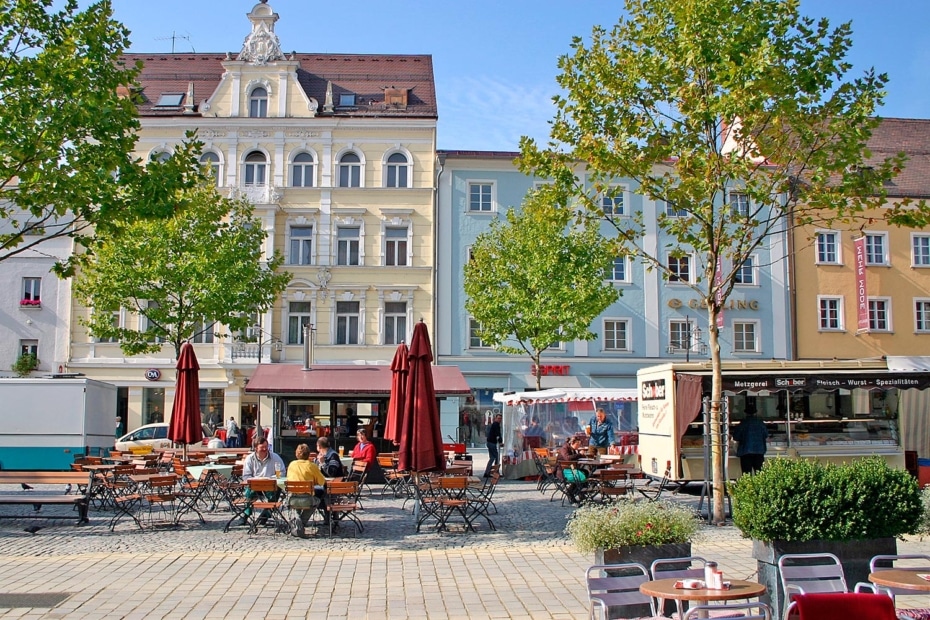 Belebter innerstädtischer Platz mit Außengastronomie, roten Sonnenschirmen, Bäumen und historischen sowie farbigen Hausfassaden.