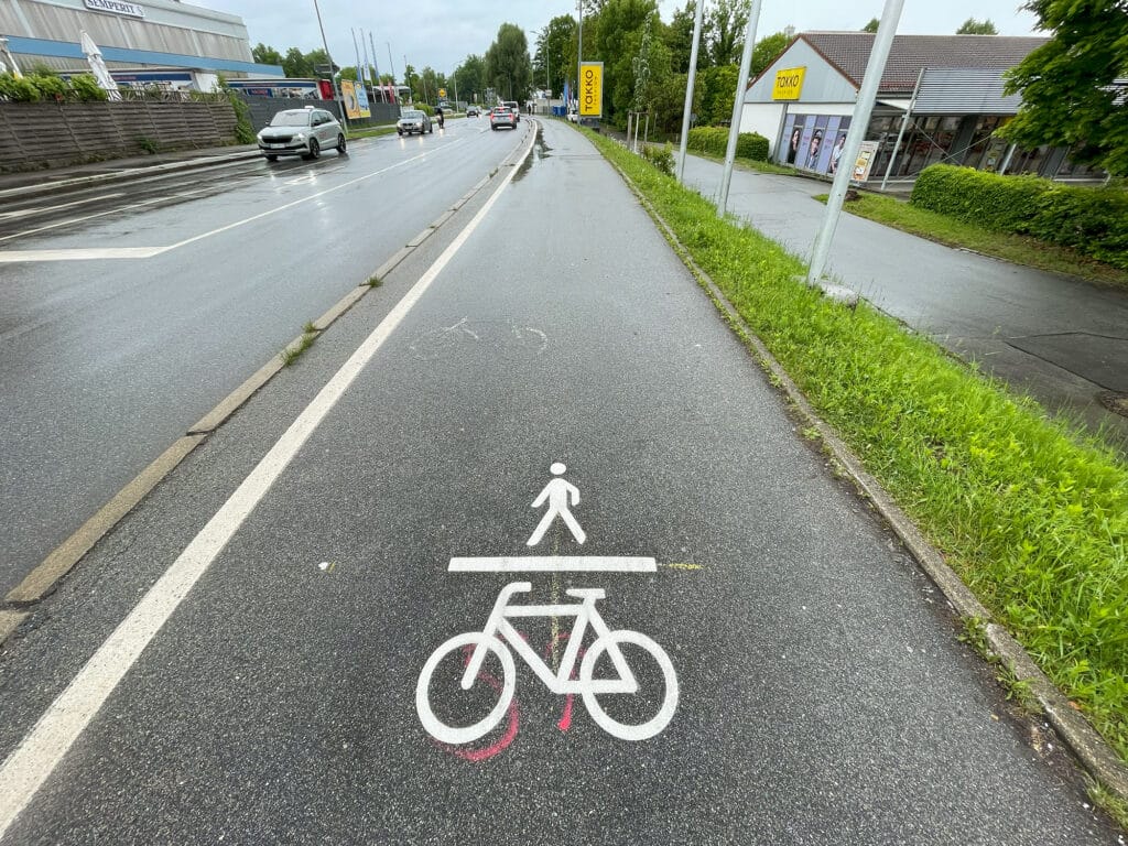 Auf einem gemeinsamen Geh- und Radweg an der Graflinger Straße sind weiße Piktogramme für Fußgänger und Fahrräder auf dem nassen Asphalt markiert.