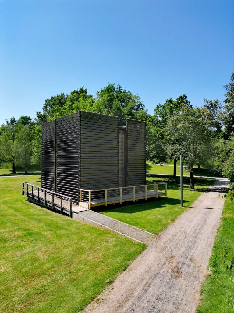 Kleiner, dunkel verkleideter Pavillon mit umlaufender Holzterrasse in einer gepflegten Parkanlage