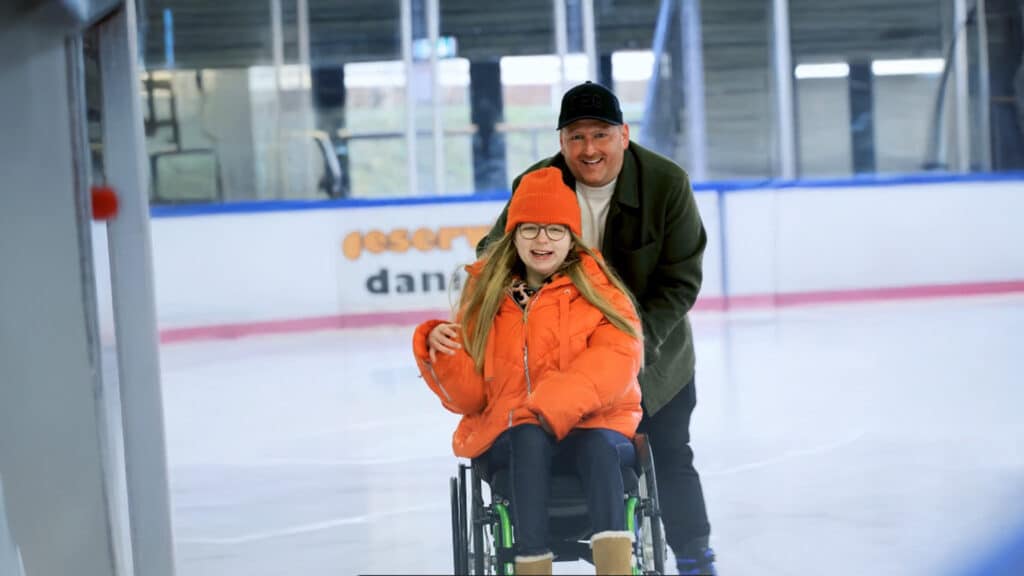 Eine junge Rollstuhlfahrerin in orangefarbener Jacke fährt auf einer Eisbahn, während ein Mann sie von hinten schiebt.
