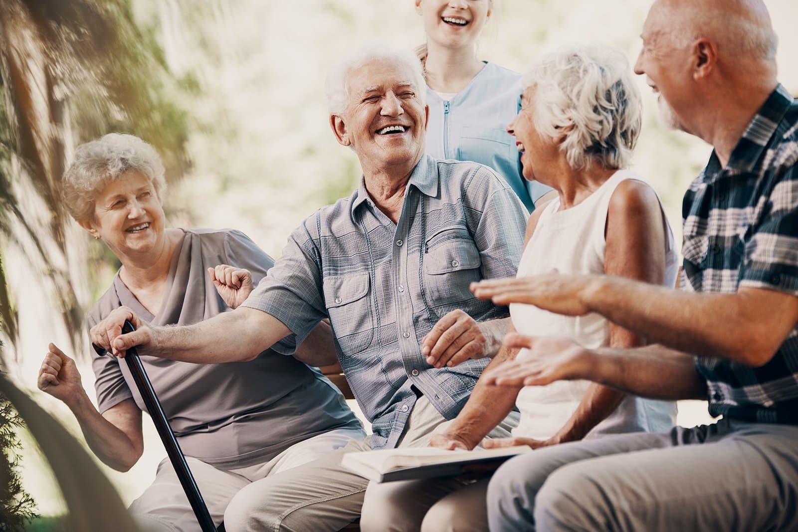 Mehrere ältere Menschen sitzen und stehen in geselliger Runde im Freien; eine Seniorin hält einen Gehstock.