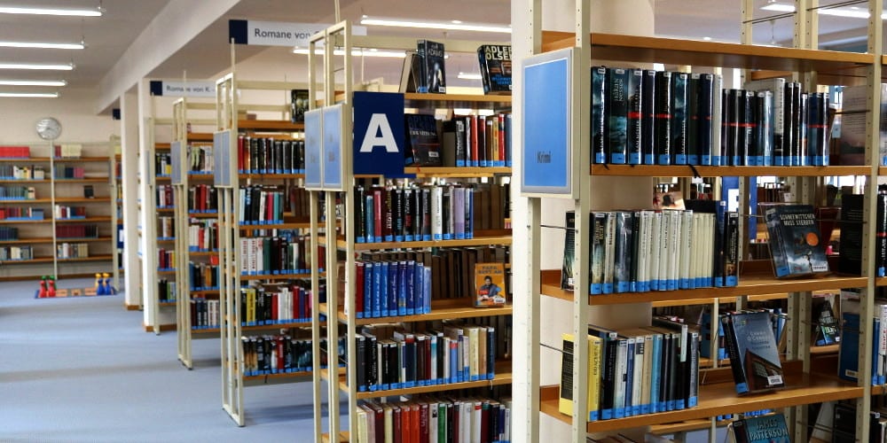 Blick durch einen Bibliotheksraum mit mehreren Reihen voller Bücherregale und einem großen Buchstaben A als Bereichsmarkierung.