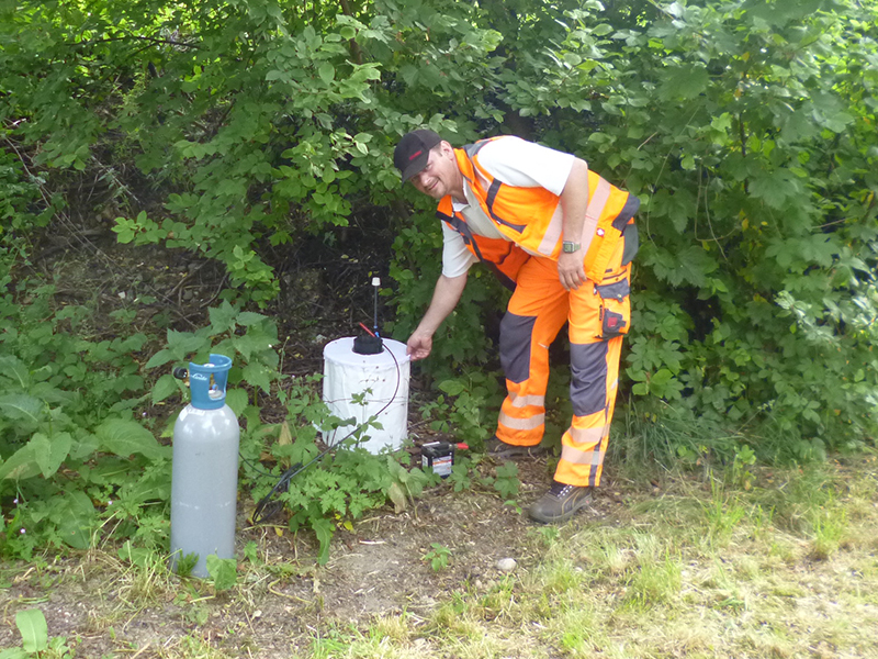Mitarbeiter in orangefarbener Warnschutzkleidung bedient am Waldrand ein weißes Gerät neben einer Gasflasche.