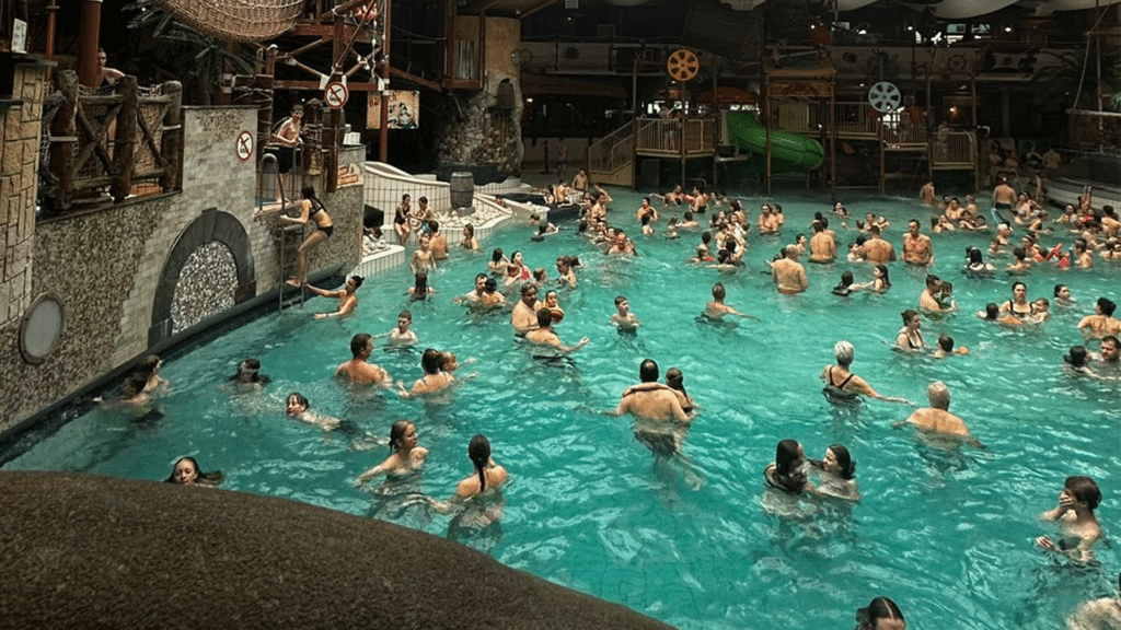 Belebtes tropisches Erlebnisbad mit zahlreichen badenden Menschen in einem großen türkisfarbenen Innenbecken, umgeben von künstlicher Felslandschaft und tropischer Dekoration.