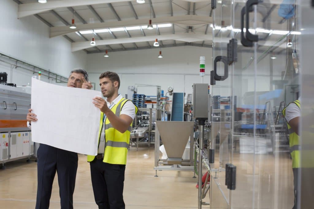 Zwei Mitarbeiter stehen in einer industriellen Produktionshalle und halten gemeinsam einen großen Plan vor eine Maschine.