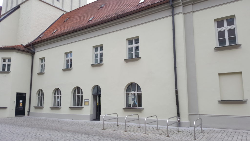 Historisches Gebäude mit hell verputzter Fassade, roten Ziegeln, Rundbogenfenstern und Fahrradständern vor dem Eingang in Deggendorf