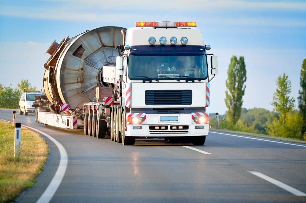 Weißer Schwerlasttransporter mit großem zylindrischem Bauteil fährt auf einer Landstraße.