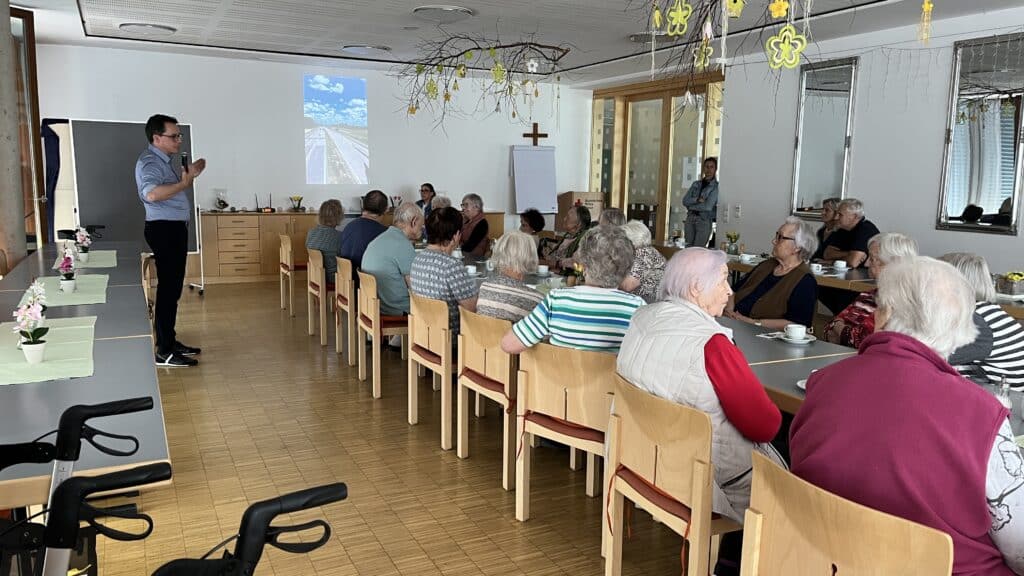 Seniorinnen und Senioren sitzen bei einer Informationsveranstaltung an Tischen in einem Saal; vorne rechts spricht eine Person mit Mikrofon, im Hintergrund ist eine Präsentation zu sehen.