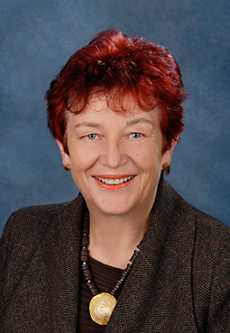 Porträt von Cornelia Wohlhüter Porträt einer lächelnden Frau mit kurzem roten Haar, dunklem Blazer und auffälligem goldfarbenem Anhänger vor blauem Hintergrund.