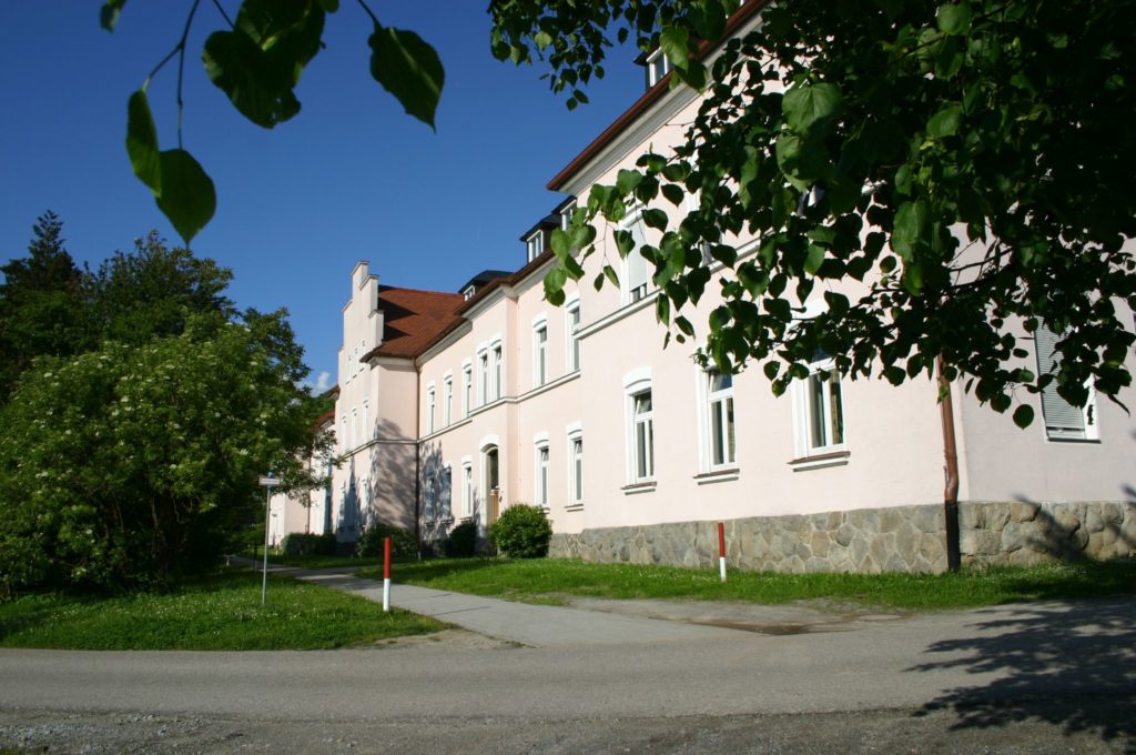 Straßenansicht eines hell verputzten, zweigeschossigen Gebäudes mit roten Ziegeldächern und Natursteinsockel, umgeben von Bäumen und Rasenflächen.