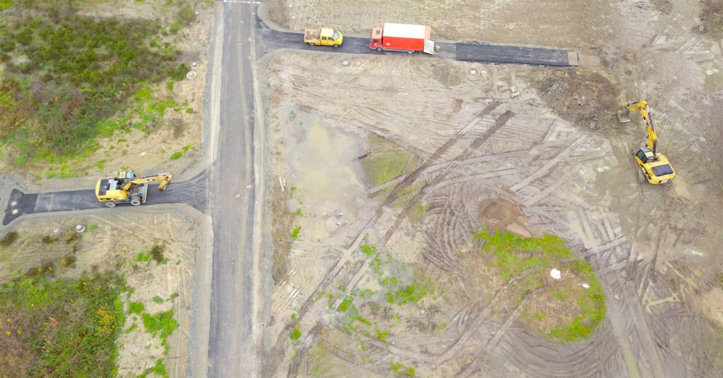 Luftaufnahme einer Baustelle mit mehreren Baufahrzeugen, neu angelegten Straßenabschnitten und unbefestigtem Erdreich.